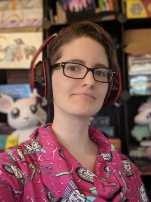 A bust-up photo of a white nonbinary person with short dark-blond hair and glasses. They are wearing a pink shirt and headphones. They are slightly smiling at the camera and sitting in front of shelves full of boardgames.
